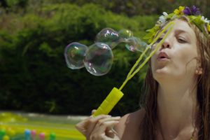 Fotograma del Videoclip de música electrónica: Chica soplando pompas de jabón en una pool party.