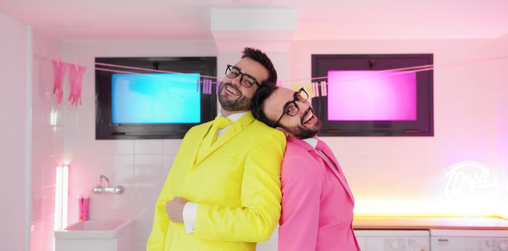 En esta toma de media distancia, se muestra a los cantantes de Tú Peleas Como Una Vaca inclinados uno sobre el otro. Uno de ellos lleva un traje completamente rosa, mientras que el otro viste un traje amarillo. La imagen captura la complicidad y la actitud relajada de los artistas, quienes transmiten su estilo y personalidad únicos a través de su vestuario llamativo y colorido. Esta escena forma parte del videoclip de "Vivir Así Es Morir de Amor", en el que se destaca la esencia nerd y friki de la banda, combinando performance y coreografía con un entorno aséptico y surrealista. Esta imagen refleja la diversión y el carácter entrañable del videoclip, creando una conexión visual con la propuesta artística de Tú Peleas Como Una Vaca.