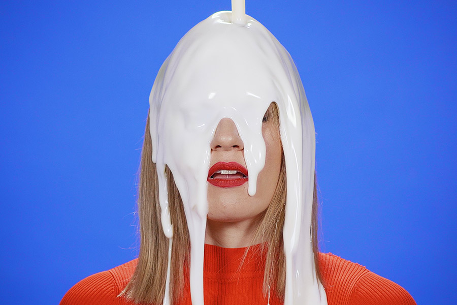 Videoclip vibrante de la canción 'Dueles' de Tú Peleas Como Una Vaca. Actriz rubia con camiseta roja y labios rojos mira intensamente a la cámara mientras cae pintura blanca, cubriendo su rostro.