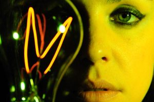 Primer plano de la actriz principal en 'Sinestesia', un cortometraje experimental producido por Colirio Films. Una bombilla vintage ilumina su cara y se refleja en su ojo derecho, aportando un brillo especial a su mirada verde. El tono global de la imagen es de un verde amarillento.