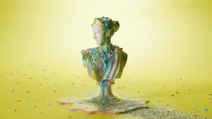 Imagen del fotograma: Estatua de mujer griega cubierta de pintura y chispas de colores sobre fondo amarillo en los vídeos de bodegón 'Bodegones De Cuarentena' de Colirio Films, productora audiovisual Madrid